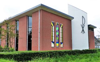 Een modern kerkgebouw met hoge raampartijen, een glas-in-loodpartij en een witte toren met het Christus-teken erop.