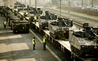 Donkergroene tanks in een rij op treinwagons. Eromheen staan militairen in camouflagekleding met gele hesjes aan.