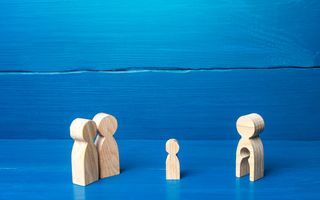 Houten figuren op blauwgeschilderde houten planken beelden relatie tussen kind, draagmoeder en ouders uit: twee grote bij elkaar, een grote apart en een kleintje ertussen.