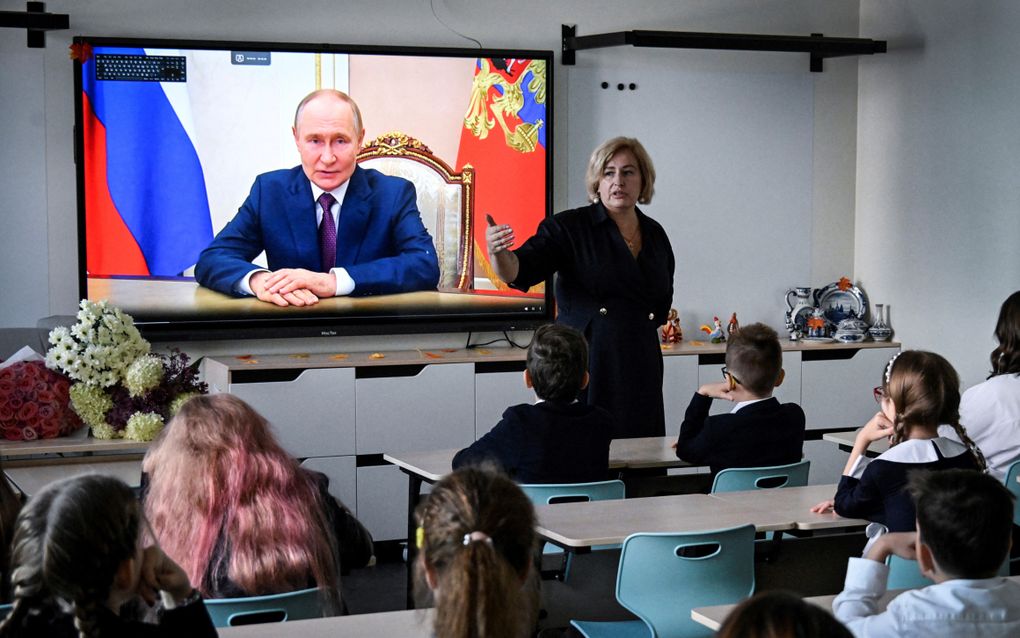 Kinderen kijken naar een digibord met daarop een man in pak op het scherm.