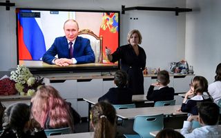 Kinderen kijken naar een digibord met daarop een man in pak op het scherm. 