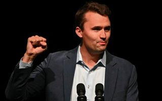 Een man met bruin haar staat voor een microfoon met een opgestoken vuist om zijn boodschap kracht bij te zetten. Hij draagt een lichtblauw overhemd met daarover een donker colbert.