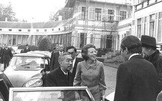 Kleine man met snor steekt hand uit naast auto.