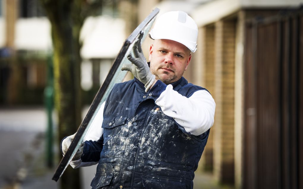 Een man in een blauwe, met stopverf besmeurde bodywarmer tilt een dikke glasplaat. Hij heeft een witte helm op en draagt grijze handschoenen. 