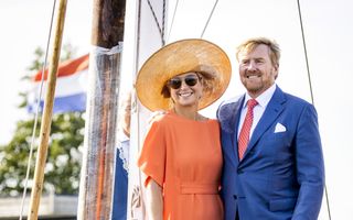 Een vrouw in een oranje jurk en hoed staat naast een man in een blauw pak op een zeilschip.
