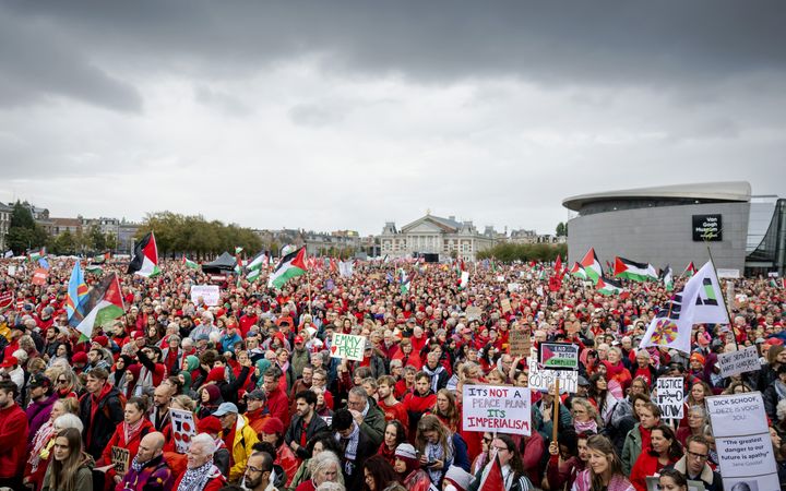 Een roodgekleurde menigte demonstreert op het Museumplein onder een grauwe hemel, met borden en vlaggen in de hand.