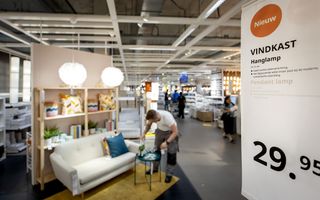 Interieur van woonwinkel IKEA. Rechts een banner met de cijfers 29,95, prijs van een hanglamp. In het midden kijkt een man met wit shirt en grijze broek naar een tafeltje op een geel vloerkleed, bank met kast staat erachter.