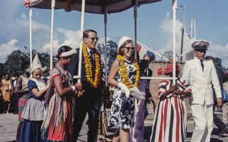 Prins en prinses lopen met oranje boa om de nek onder een soort tent die omhoog wordt gehouden door Surinamers.