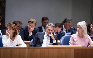 Een man met pak met naast hem twee vrouwen. Achter de man zitten nog meer mannen in pak. 