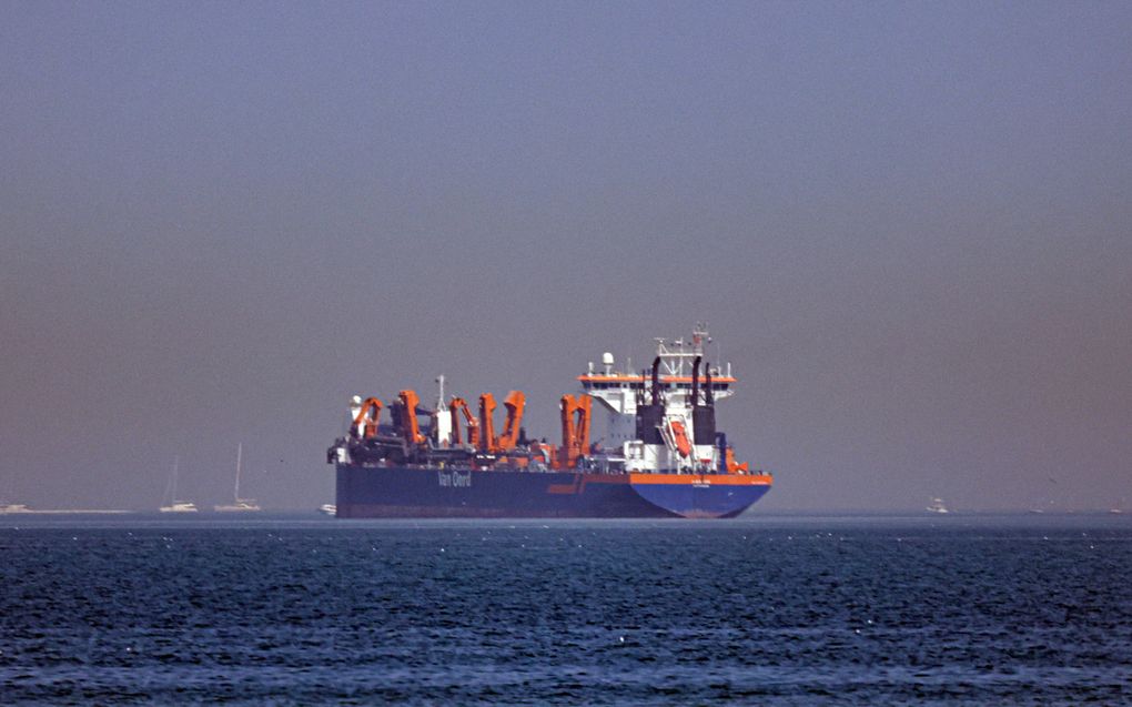 Een blauw schip met rode installaties ligt op zee. Op de achtergrond is de kust te zien. Op de zijkant van het schip staat Van Oord.