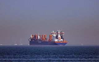 Een blauw schip met rode installaties ligt op zee. Op de achtergrond is de kust te zien. Op de zijkant van het schip staat Van Oord.