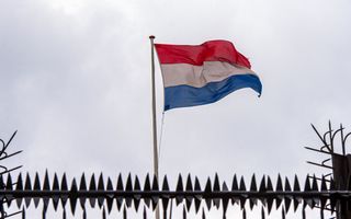 Een Nederlandse vlag wappert in de wind aan een vlaggenmast achter een metalen hek met scherpe punten en haken.