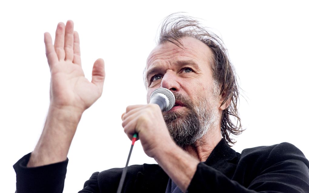 Een wat oudere man met grijzend baardje en lang haar houdt een microfoon vast. Zijn rechterhand houdt hij in de lucht.