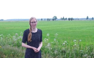 Een jonge vrouw, met bril, haar in staart, donkere jurk. Achter haar een weids graslandschap, daarachter heuvels.