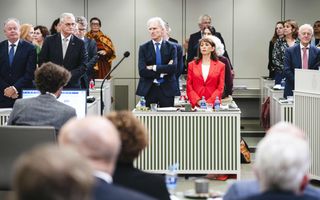 Mannen in pak staan achter witgroene bureautjes. Ook staan er vrouwen naast hen. Een vrouw heeft een rode blazer aan. 