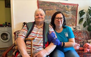 Een oude vrouw zit op een bank, met naast haar een jongere vrouw die haar arm vasthoudt. Achter hen hangt een wandkleed.