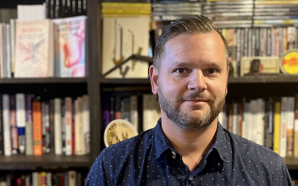 Man, dertiger, in blauw overhemd voor boekenkast.