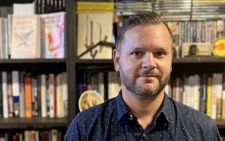 Man, dertiger, in blauw overhemd voor boekenkast.