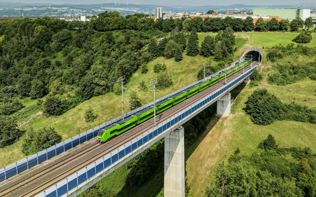 Luchtfoto van een groene hogesnelheidstrein die door een heuvelachtig landschap rijdt. Op de achtergrond is bebouwing te zien.