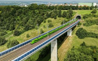 Luchtfoto van een groene hogesnelheidstrein die door een heuvelachtig landschap rijdt. Op de achtergrond is bebouwing te zien. 
