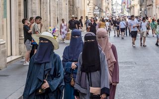 Vier vrouwen in blauwe en roze lange gewaden die tot de grond raken, lopen over een straat. Alleen de ogen van de vrouwen zijn zichtbaar.