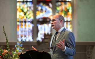 Man in groengrijs pak met bril op staat achter spreekgestoelte.