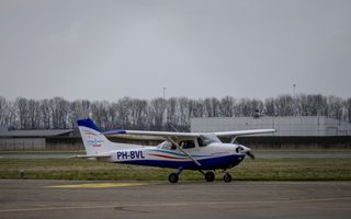 Een klein vliegtuigje, in de kleuren blauw, wit en rood. Het staat op de grond, erachter gebouwen van Lelystad Airport.