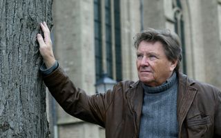 Een oudere man met grijzend haar poseert voor een grote oude kerk. Hij draagt een blauwe koltrui en een bruine leren jas, en leunt met een hand tegen een boom.