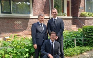 Drie mannen van middelbare leeftijd poseren in een zwart pak voor een jarendertigwoning.