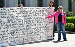 Een vrouw met een roze jas wijst naar een enorme banner met daarop namen van jonge slachtoffers.