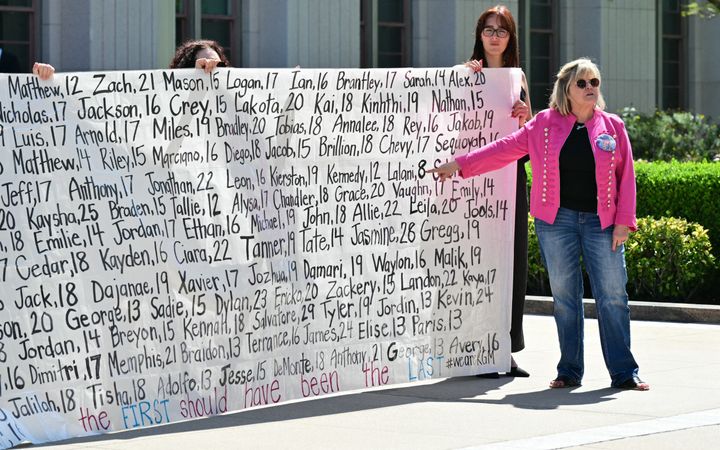 Een vrouw met een roze jas wijst naar een enorme banner met daarop namen van jonge slachtoffers.