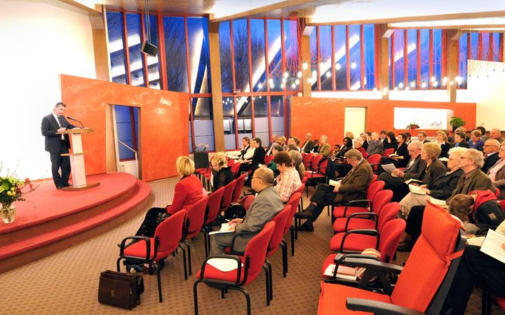 Een zaal met mensen op rode stoelen; achter een katheder staat een man. 