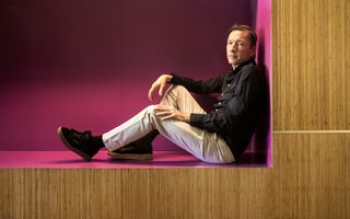 Man van middelbare leeftijd met lichte broek en donkerblauw overhemd zit in een grote nis waarvan de wanden roze geverfd zijn. De nis zit in een wand die met houtstructuren is bedekt.