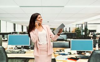 Vrouw in lichtroze pakje staat voor een bureau. Ze heeft een telefoon aan haar oor en een A4’tje  in haar hand. Ze lacht. In de kantoorruimte zijn meerdere bureaus met computerschermen.