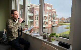 Een man zit voor de vensterbank en houdt een kaart met vogelsoorten en een verrekijker vast, terwijl hij uit het raam tuurt.