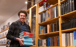 Een man met donker krullend haar houdt een stapel blauwe en rode boeken vast. Achter hem staat een lichte eiken kast vol boeken. 