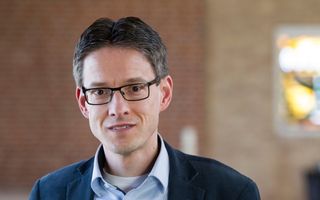 Man met donker haar, bril met rechthoekig montuur, bruine ogen, lichtblauw overhemd en donkerblauw jasje. 