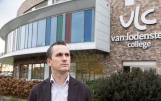 Man met gestreept overhemd en colbert staat voor een eigentijds schoolgebouw met grote raampartij.