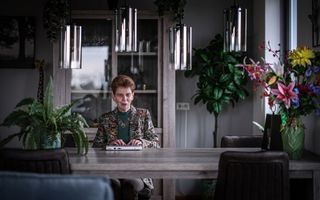 Vrouw aan een tafel met veel groen en een vaas met kleurige bloemen om zich heen.