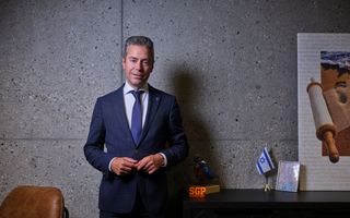 Man met blauw pak en grijzend haar staat naast een kastje met een vogeltje en een vlaggetje van Israël daarop.