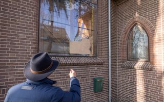 Een man met een hoed wijst naar een glas-in-loodraam waar een barst in zit.