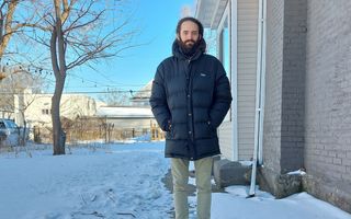 Een bebaarde man staat op een besneeuwd tuinpad naast een huis. 