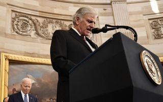 Van onderaf genomen foto van een grijze man in zwart pak die achter een zwart spreekgestoelte in een microfoon spreekt. Op de achtergrond kijkt voormalig president Joe Biden bedrukt naar de grond. 