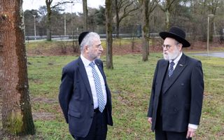 Twee mannen in pak, de een met een hoed op, staan op het gras, met op de achtergrond bomen en een weg.