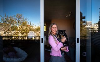 Een vrouw met een roze vestje aan staat in de opening van schuifdeuren met een hond op de arm.