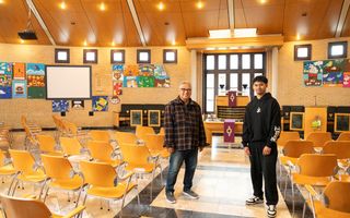 Een man met grijs haar en een bril en een jongen met zwart haar staan in het gangpad tussen rijen stoelen in een kerkzaal, met op de achtergrond een preekstoel en aan de wand kindertekeningen.