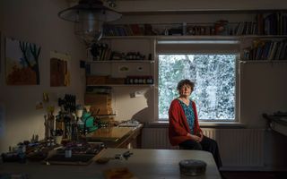 Donkere foto van een vrouw met kort, bruin haar en een rood vest aan. Ze zit op een kruk voor een raam met uitzicht op besneeuwde takken.  