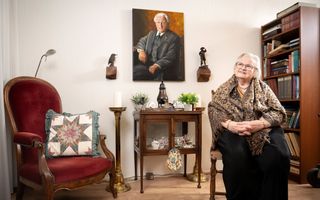 Oude vrouw met zwarte rok en bruinige doek om haar schouders zi tin een stoel, kijkt schuin naar boven. Achter haar een boekenkast.