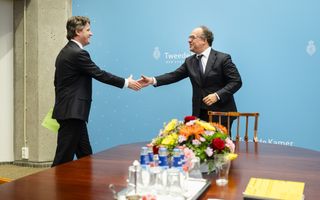 Twee mannen in pak begroeten elkaar. Op de voorgrond een ronde tafel met bloemen en een gevuld dienblad.