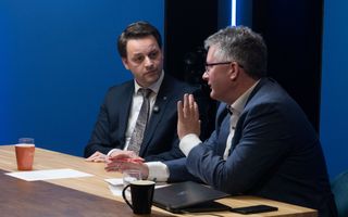 Twee mannen  in een donkerblauw pak, van middelbare leeftijd, zijn aan tafel met elkaar in gesprek.